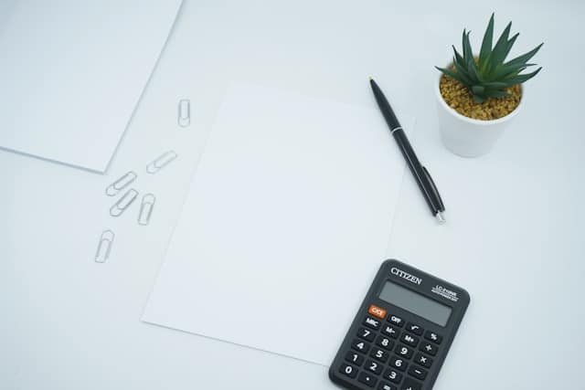 A cactus, calculator, pen, paper, and paperclips arranged on a white table.