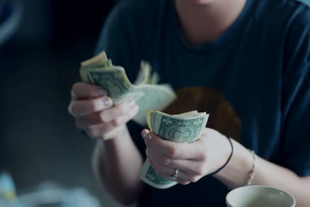 A person counting dollar bills at a table with a cup nearby 